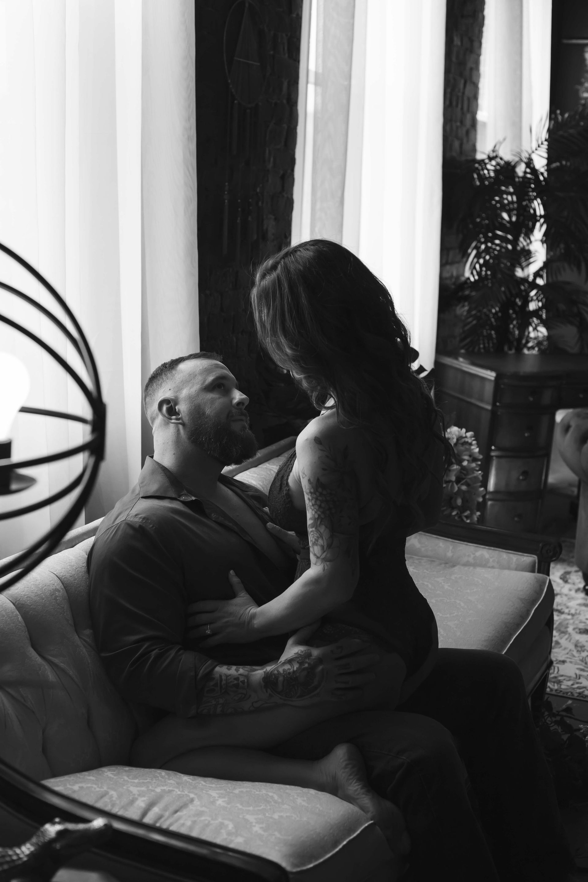 Man and woman embrace on a couch. The woman sits atop the man, looking down. Black and white, indoors.