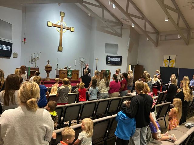A congregation stands in a church during a service, facing a large wooden cross mounted on the wall behind the altar.