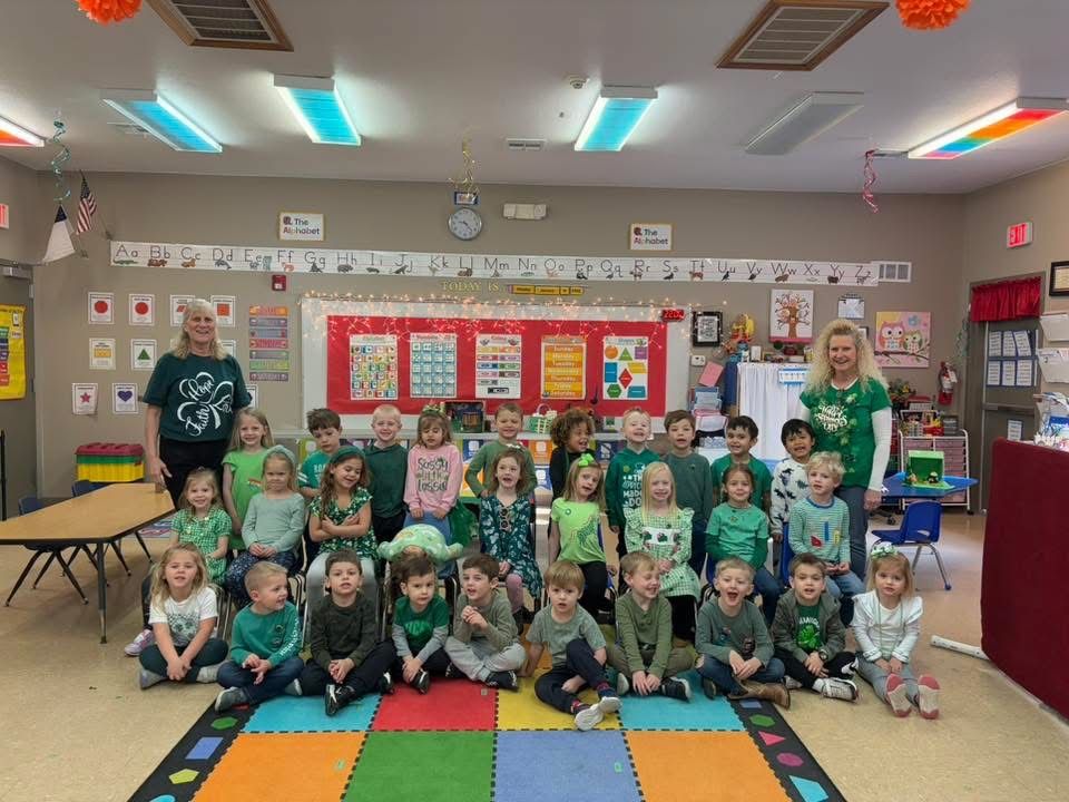 A group of children and two adults dressed in green posing for a photo in a brightly decorated classroom.
