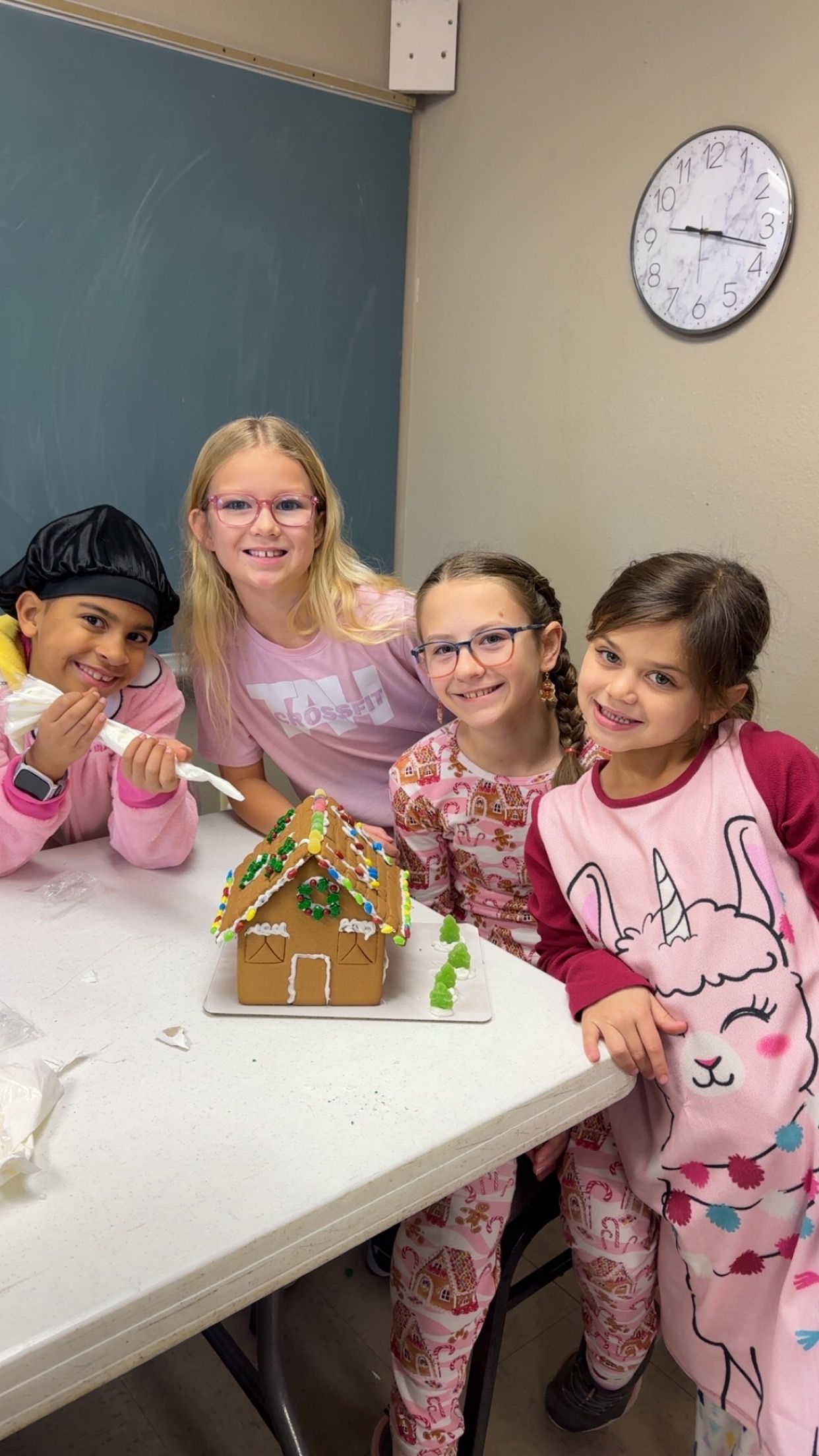 Four people in pajamas assemble a gingerbread house on a white table, smiling together in a room with a wall clock.
