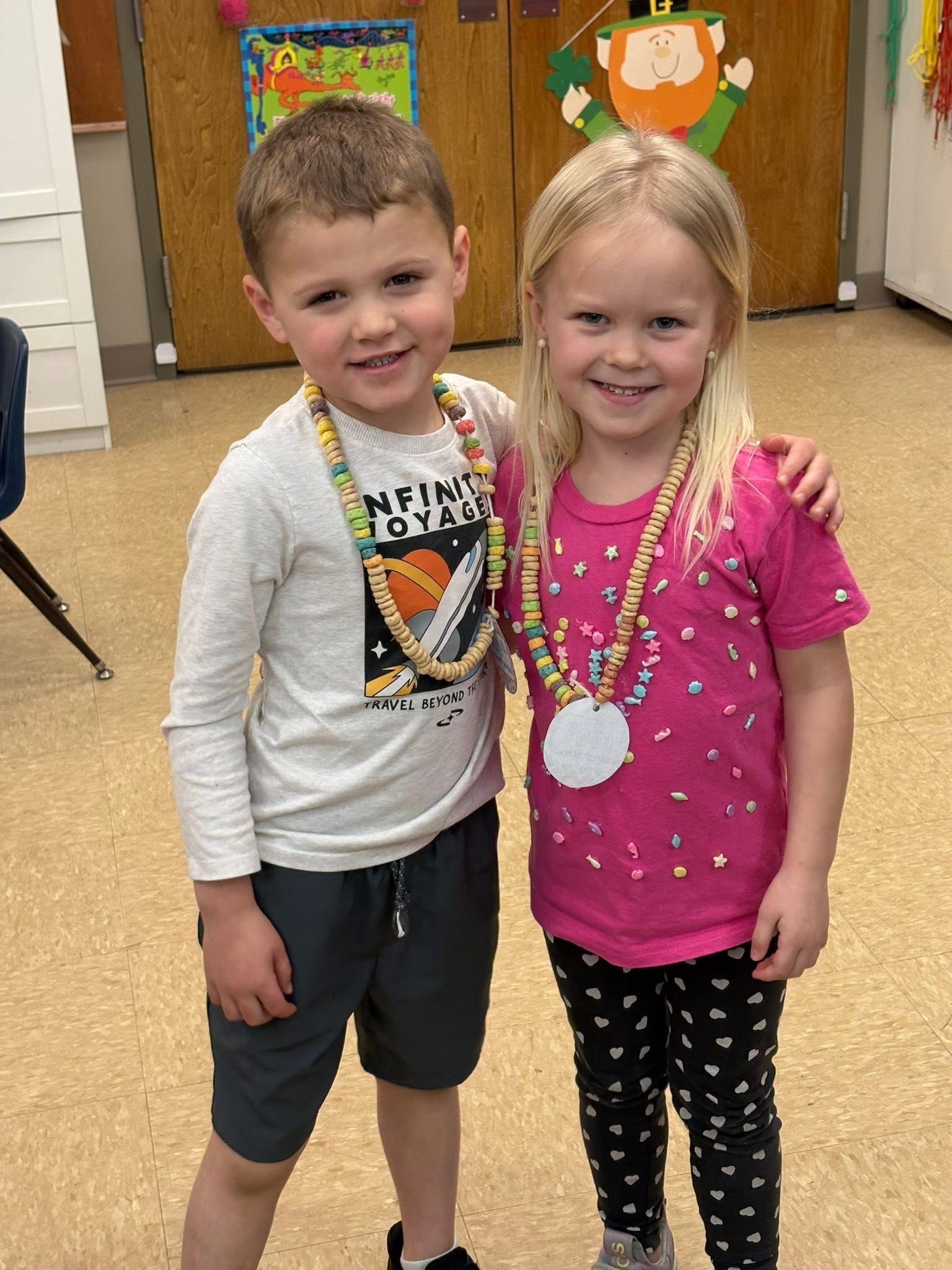 Two smiling children wear candy necklaces and casual clothes while standing in a classroom with festive wall decorations.