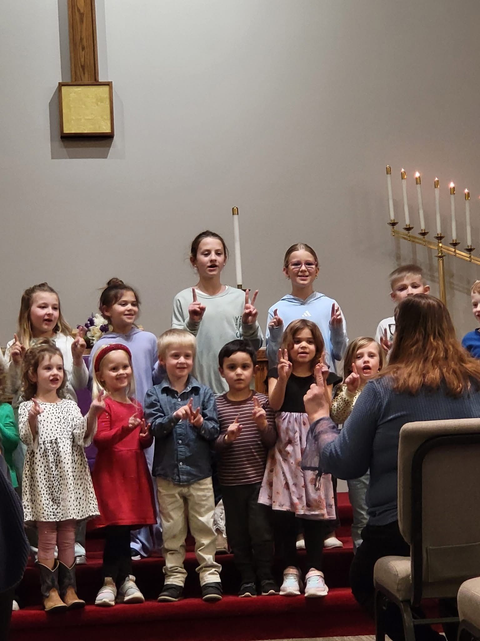 A group of children performing on a church stage, standing in rows while making hand gestures, facing an instructor.