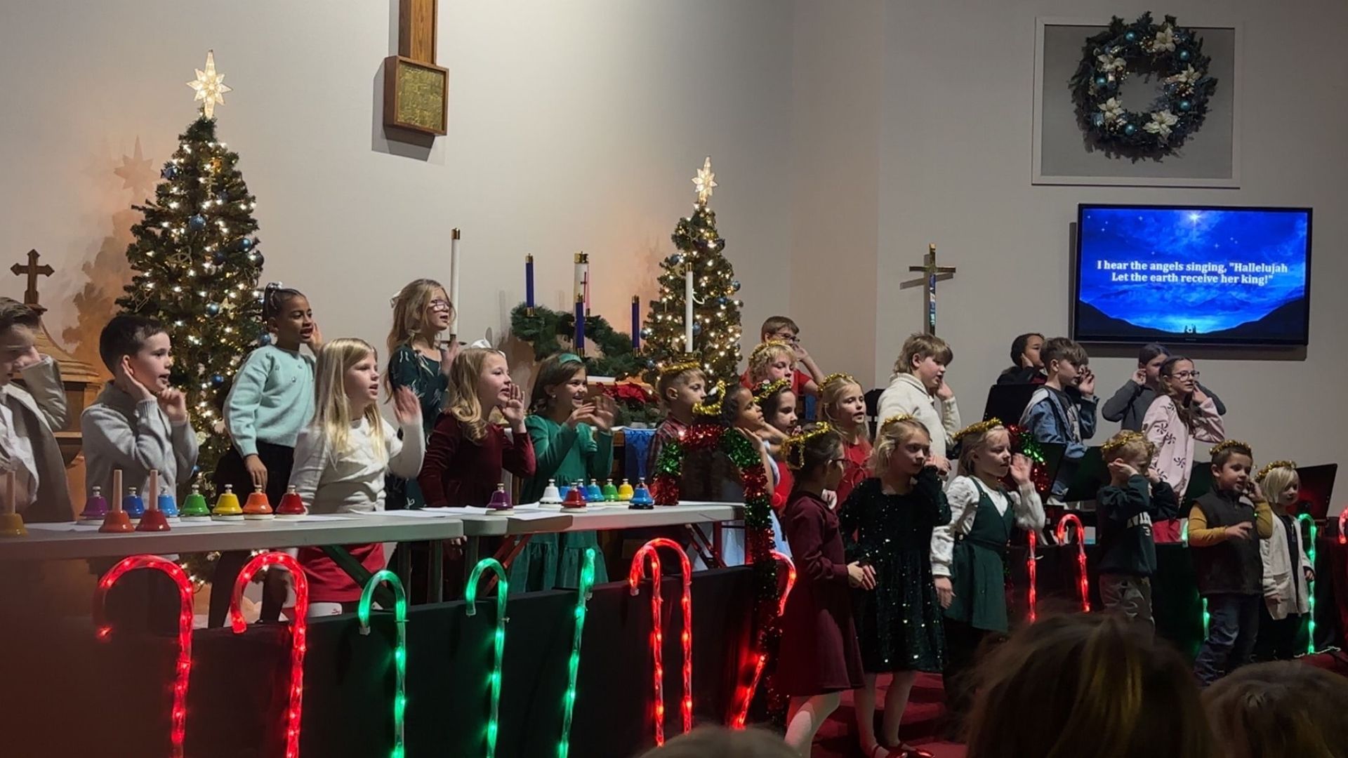 Children perform on a stage decorated with Christmas trees and illuminated candy cane lights in a church setting.