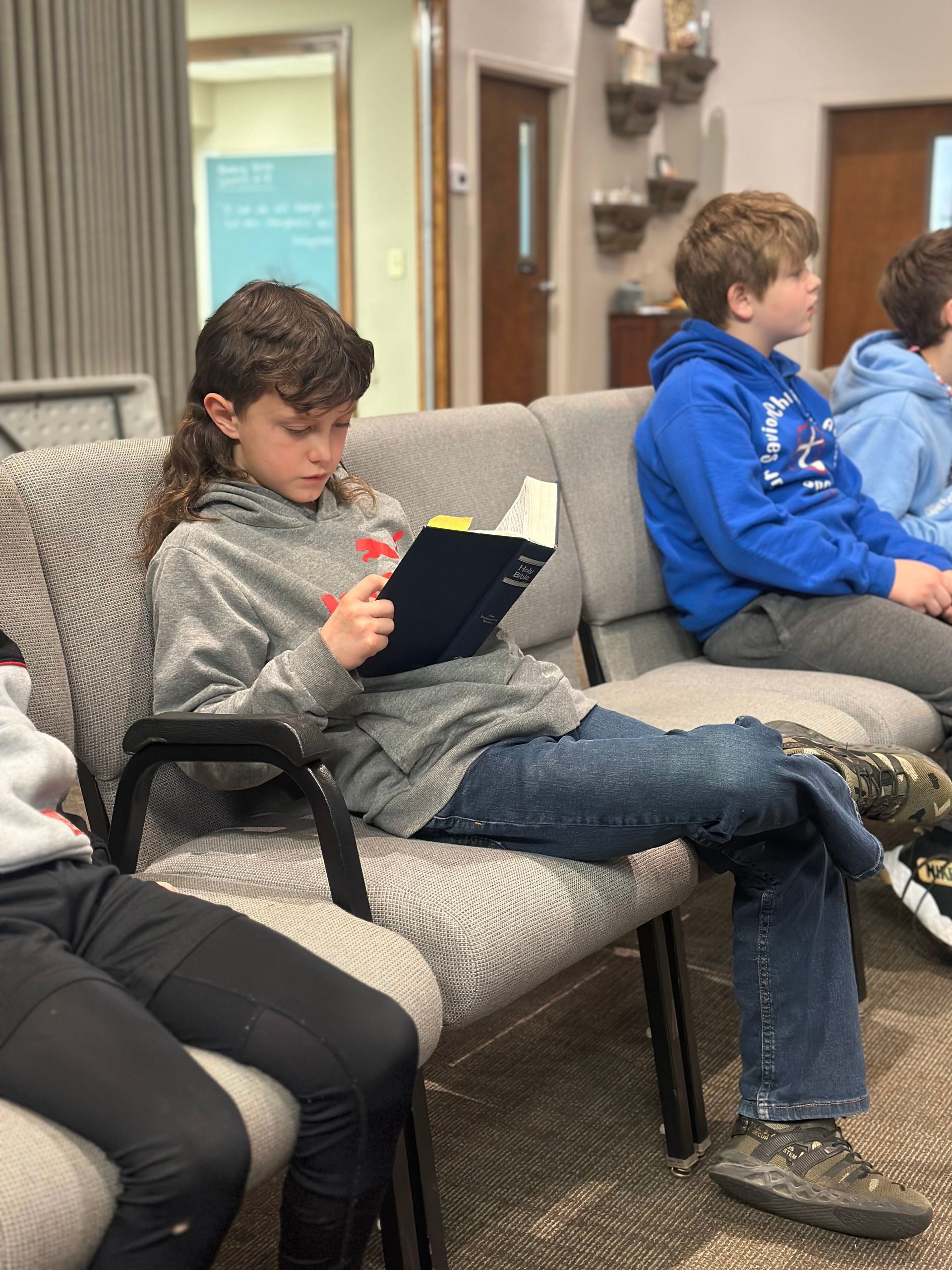 A child in a grey hoodie sits in a row of chairs reading a small, dark-covered book. Others sit nearby in a quiet setting.