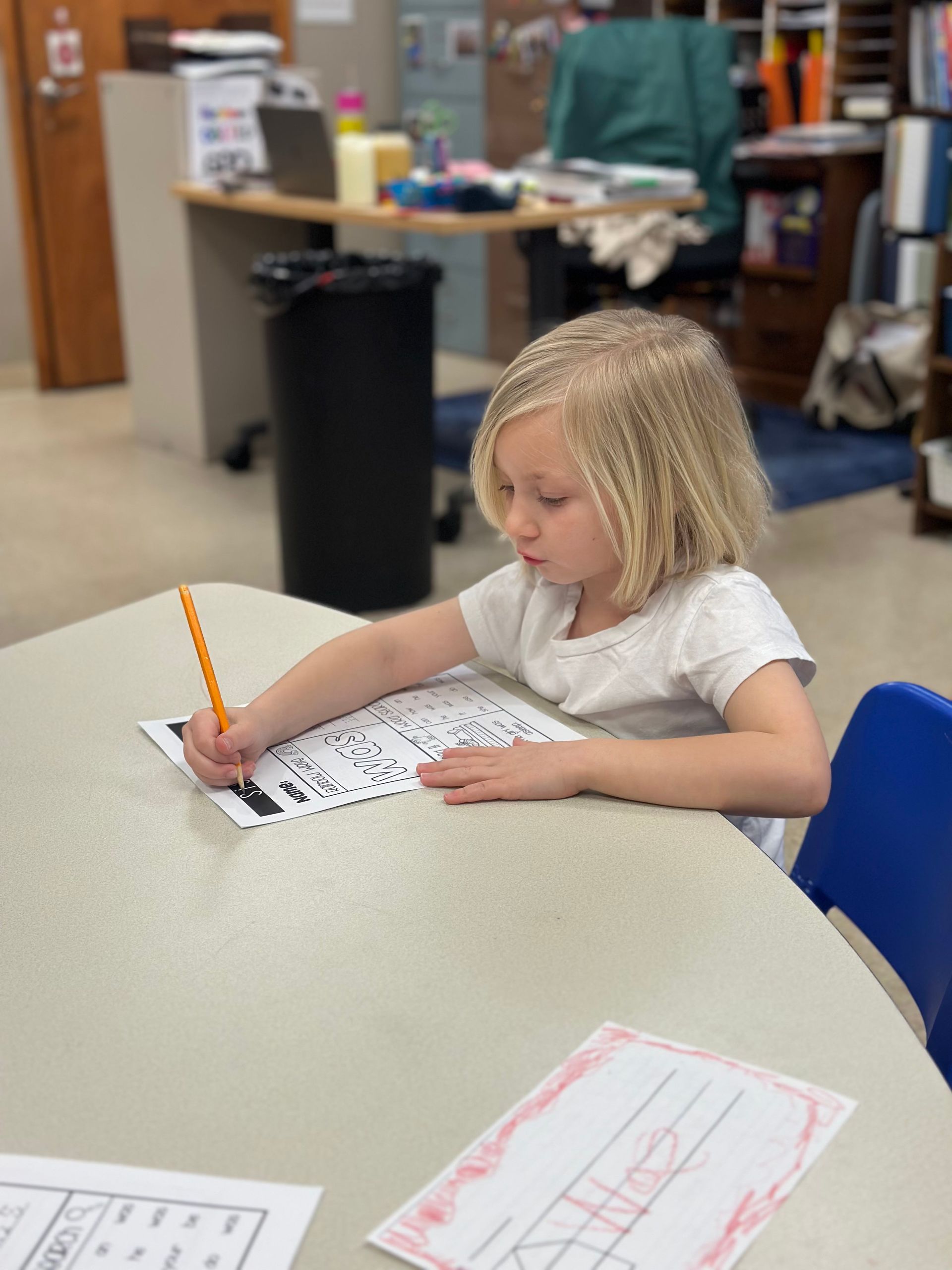 A child sits at a table in a classroom, focused on writing on a worksheet with a pencil.
