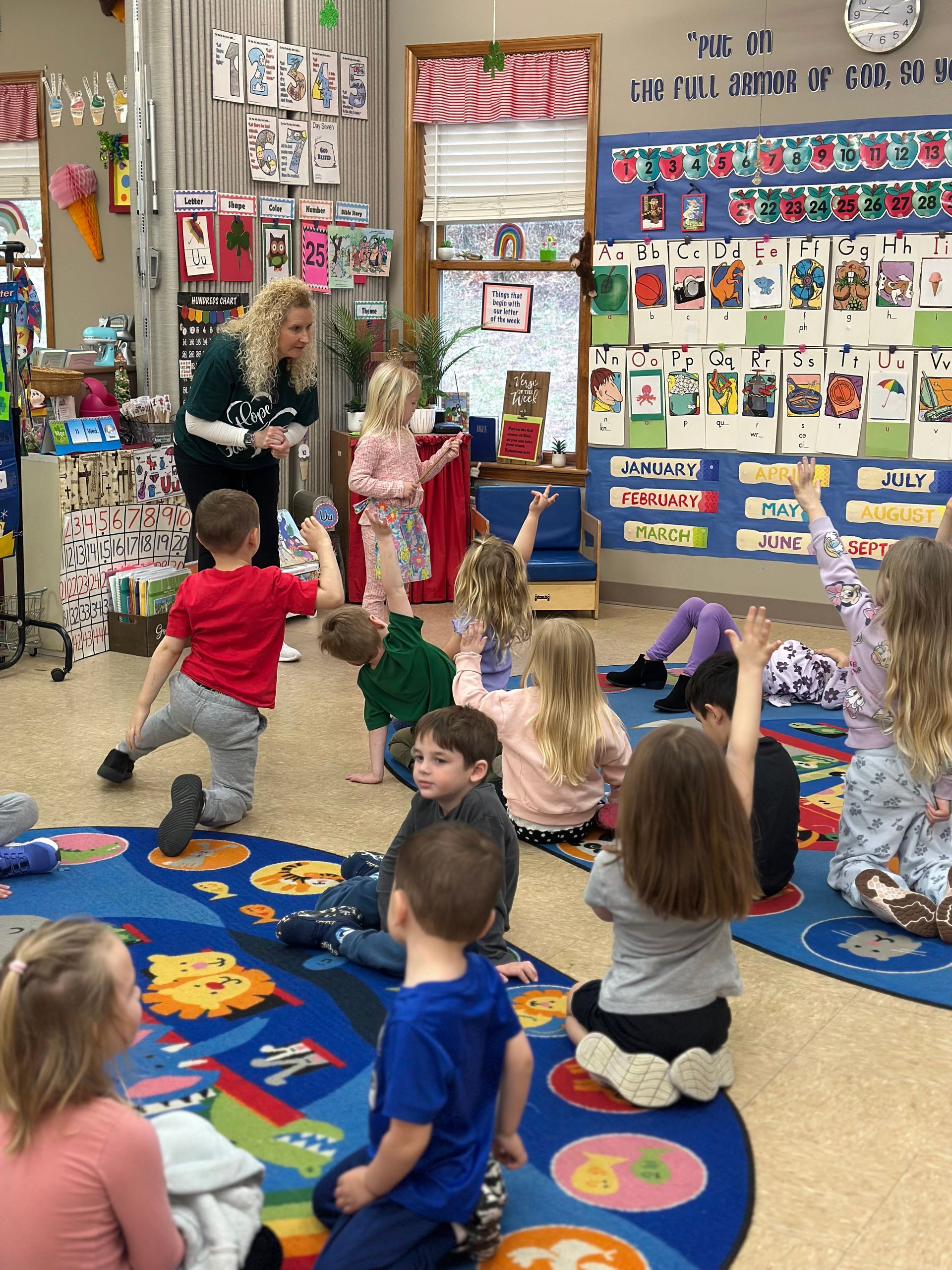 A teacher stands before a classroom of students seated on a colorful rug, with several children raising their hands.