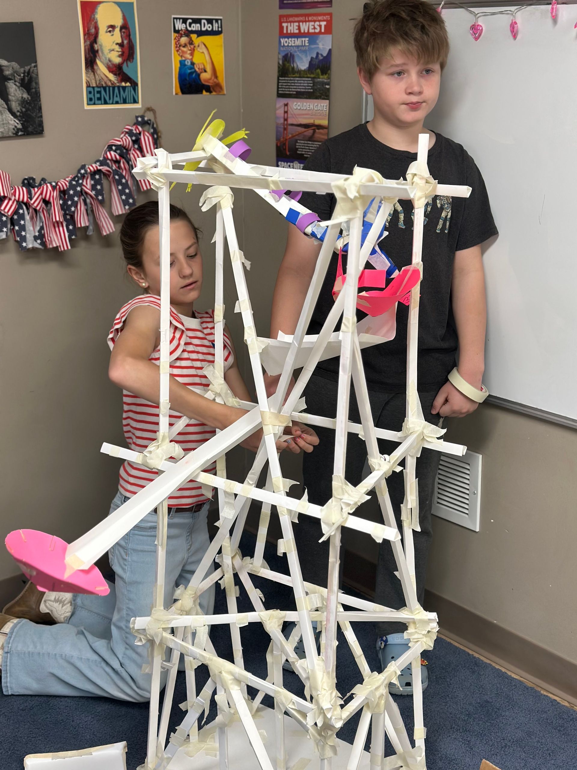 Two students stand by a tall, tape-constructed tower model with a pink platform in an indoor classroom setting.