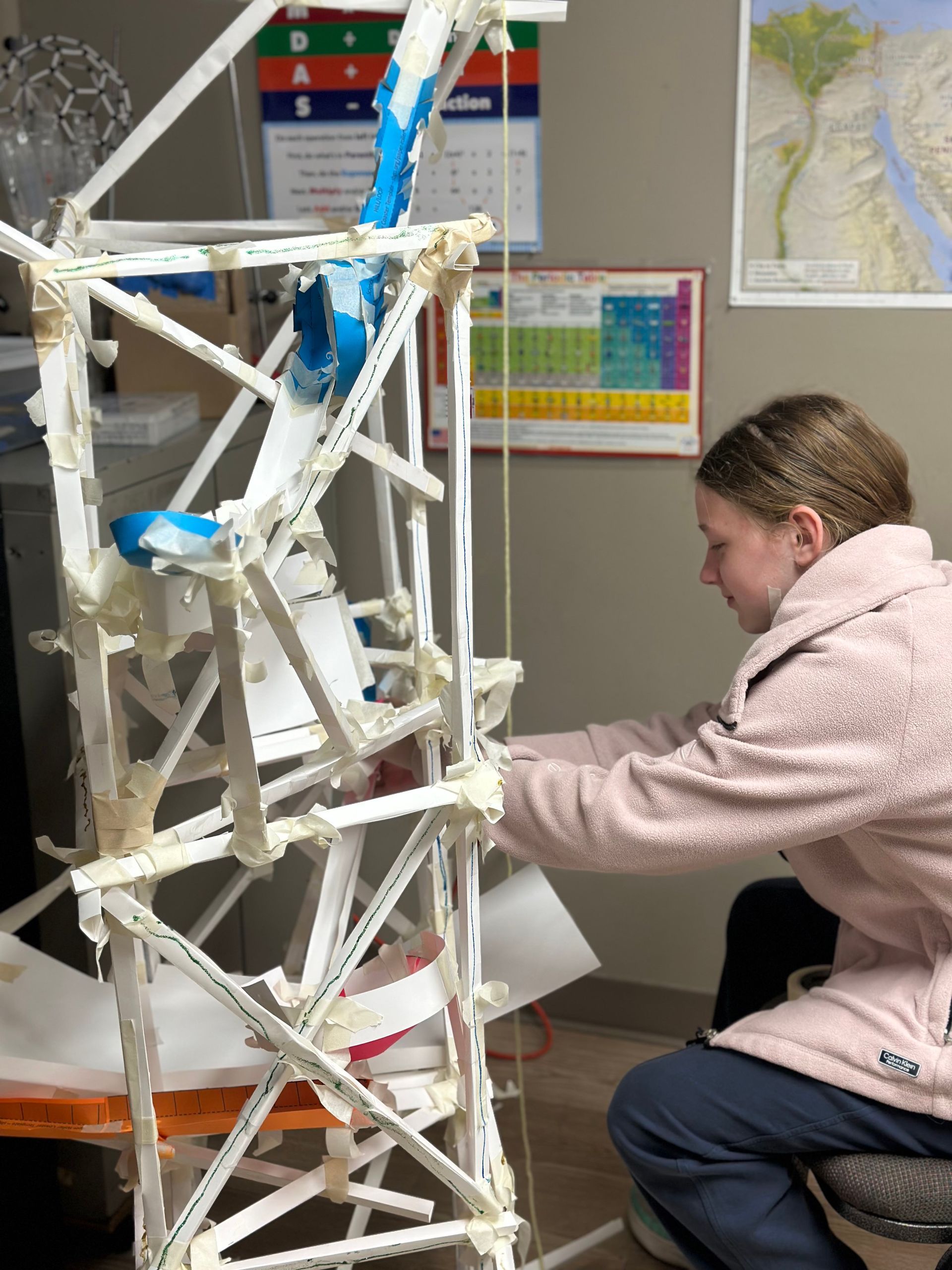 A student working on a white, tower-like sculpture made of straws and tape in a classroom setting.