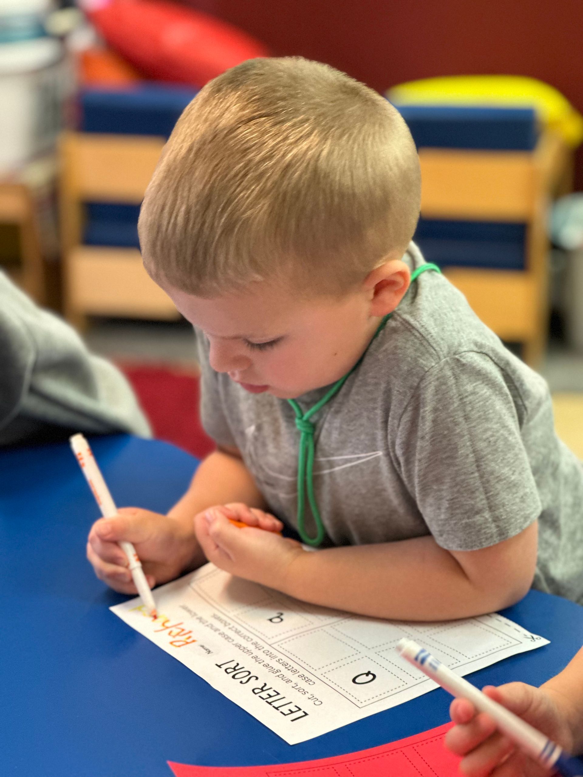 A child leans over a blue table, using a marker to complete a Letter Sort worksheet in a classroom setting.