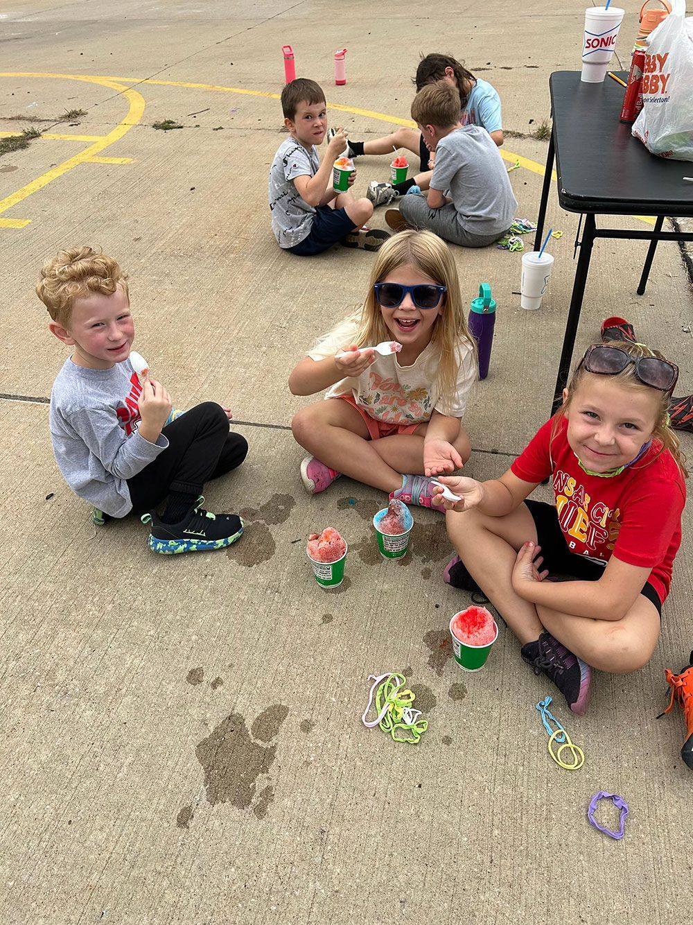 A group of children sitting on an outdoor paved surface, eating colorful shaved ice treats from small cups.