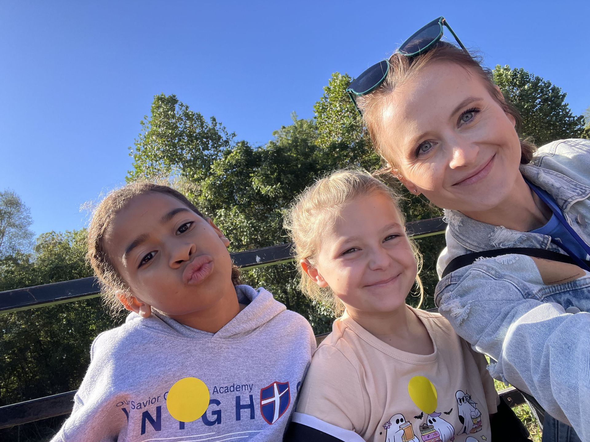 Three people smiling and posing for a selfie outdoors against a background of green trees and a clear blue sky.