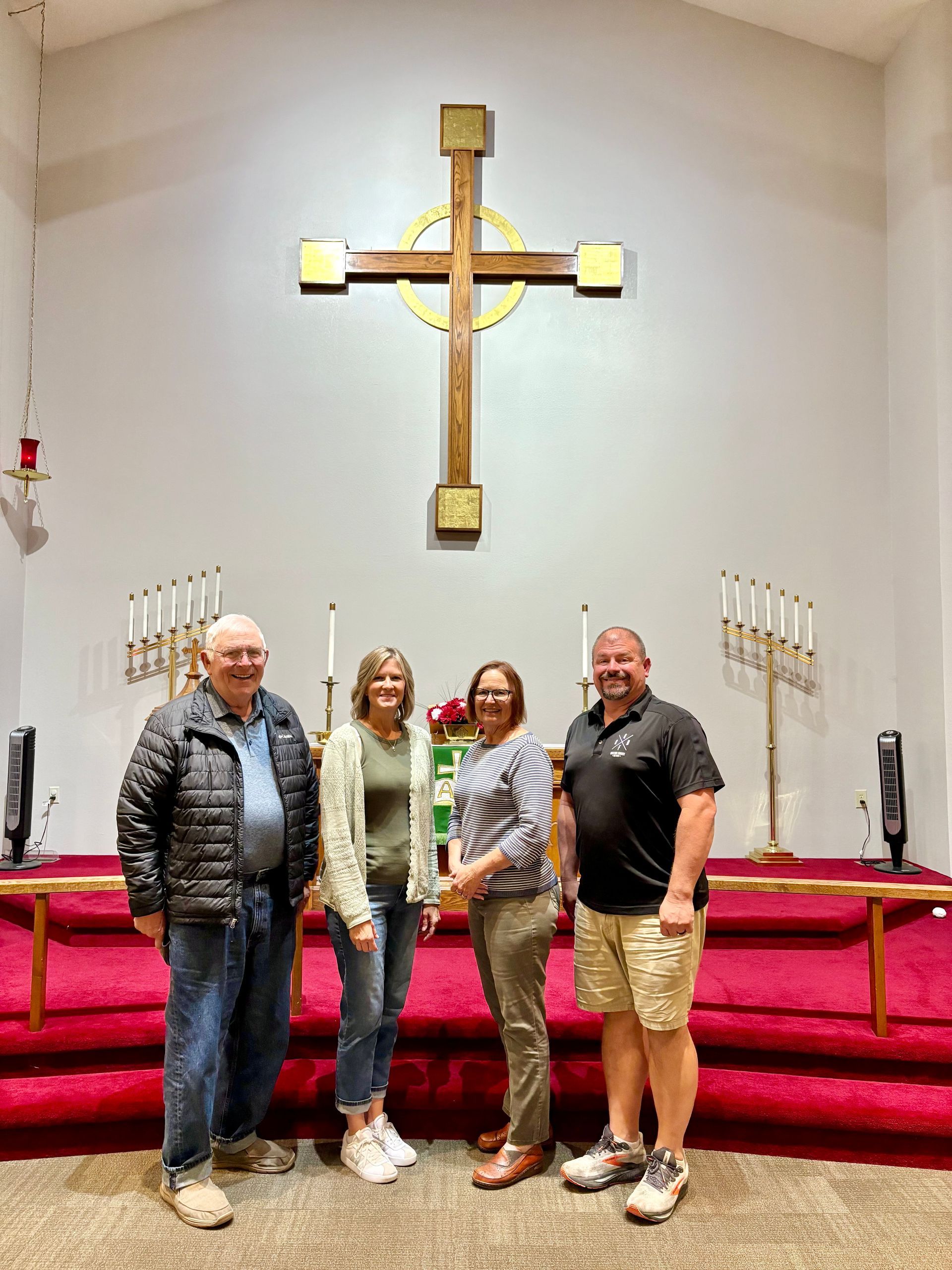 Four people stand in a line on a red-carpeted church altar in front of a large wooden Celtic cross.