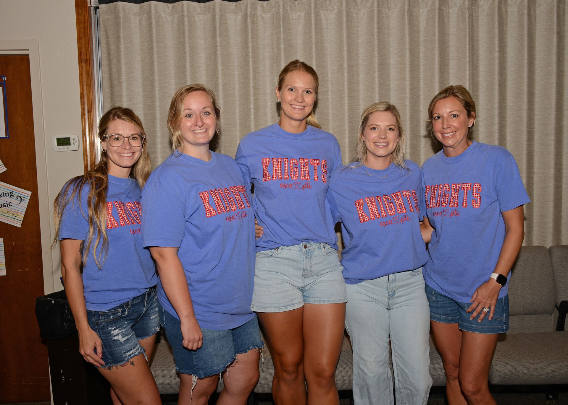 Five women in matching blue shirts and jeans posing indoors in front of beige couches.