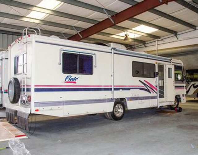 White Fleetwood RV parked indoors in a warehouse, side view with purple and pink stripes.
