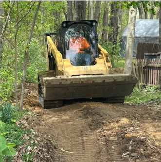 Yellow skid steer grading a dirt path in a wooded area.