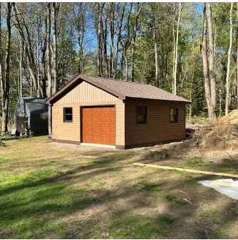 Brown garage with tan siding, door, windows, and brown roof, in a grassy area surrounded by trees.