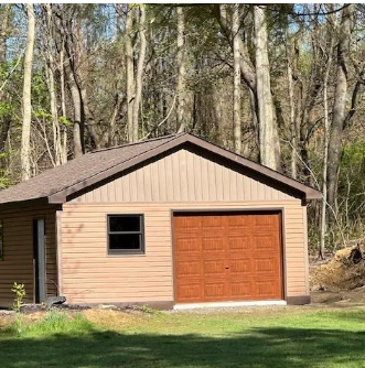 Tan garage with a brown door and roof, set against a wooded backdrop.