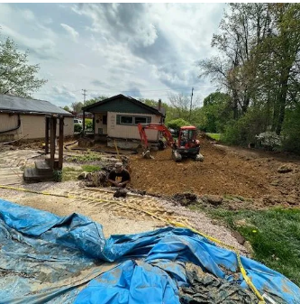 Construction site with an excavator digging near a small house; blue tarp in the foreground.