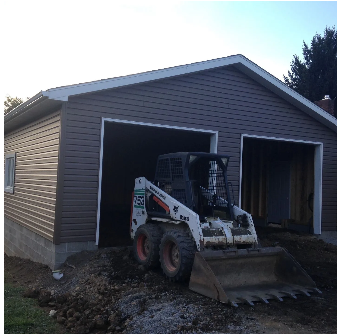 A small skid steer loader inside a two-bay garage under construction, with dark brown siding.