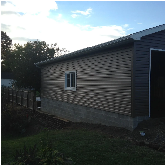 Garage with gray siding and window on a concrete foundation, in a grassy yard under a cloudy sky.
