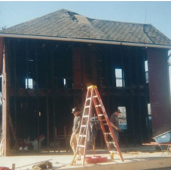 House undergoing renovation, with visible frame. Two men by a ladder, one working.