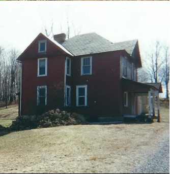 Red, two-story house with a slate roof. Small porch on the side. Unkempt yard in front.