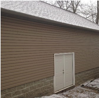 Tan siding and white double doors on a building with a snow-covered roof and brick foundation.