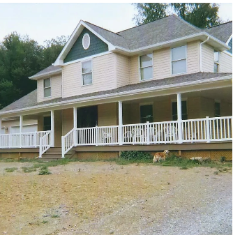 Two-story house with a large porch. Cream siding, white railings, a dog in the yard, and a gravel driveway.