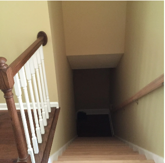 Staircase leading downwards; dark space at the bottom; wooden steps and railing; beige walls; sunlight at top.