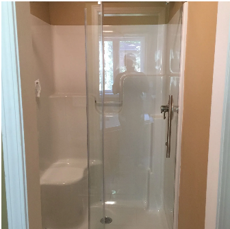 White-walled shower with a clear glass door. Includes a built-in seat and chrome fixtures. A person is reflected in the glass.