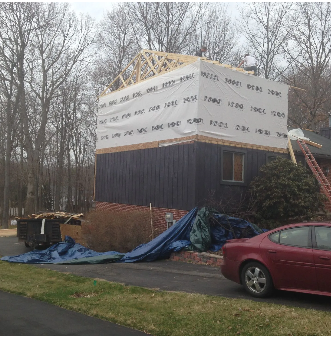 House under construction with tarp-covered upper level, wooden frame visible, near a red car and trees.