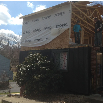 House under construction, workers on a wooden frame, partially covered in white wrap.