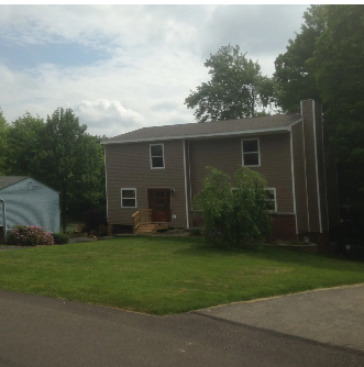 Two-story brown house with green lawn, blue shed, and trees. Cloudy sky.