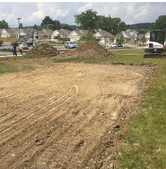 Dirt patch with tire tracks and pile of dirt; excavator and townhouses in the background.