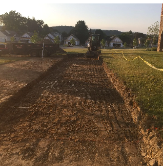 Excavation site for a pathway in a residential area. Dirt trench dug alongside a grass lawn, small excavator in background.