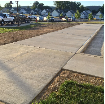 Newly poured concrete sidewalk in front of residential houses; some construction vehicles and lawn.