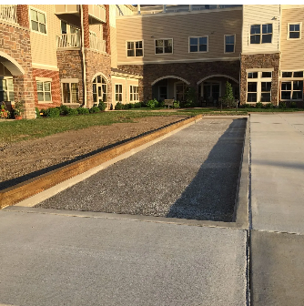 Bocce ball court with gravel surface, edged with wood, next to a paved area. Large multi-story building in background.