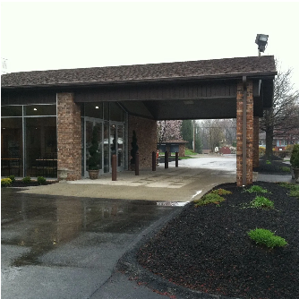 Brick building with a covered entrance, featuring glass doors. Black asphalt and mulch with greenery.
