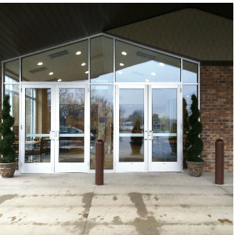 White glass doors with angled windows. Brick building with concrete entrance, flanked by potted trees.