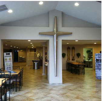 Interior of a building with a large cross on a pillar; tables and chairs in the foreground.