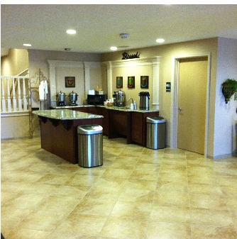 Hotel breakfast bar with coffee station, granite countertops, and stainless steel trash cans on tan tiled floor.