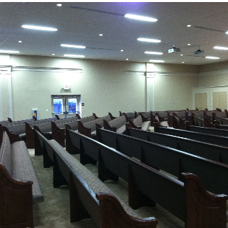 Rows of empty wooden pews in a large room with beige walls and overhead fluorescent lights.