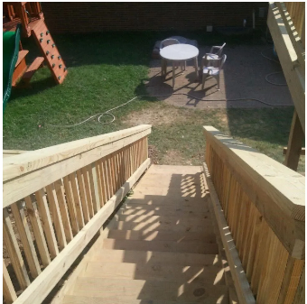 Wooden stairs leading down to a backyard with a play structure, grass, and a patio with a table and chairs.