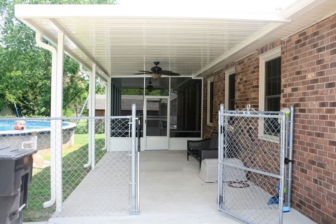 A covered brick patio features a screened enclosure, a ceiling fan, and a chain-link fenced area next to an outdoor pool.