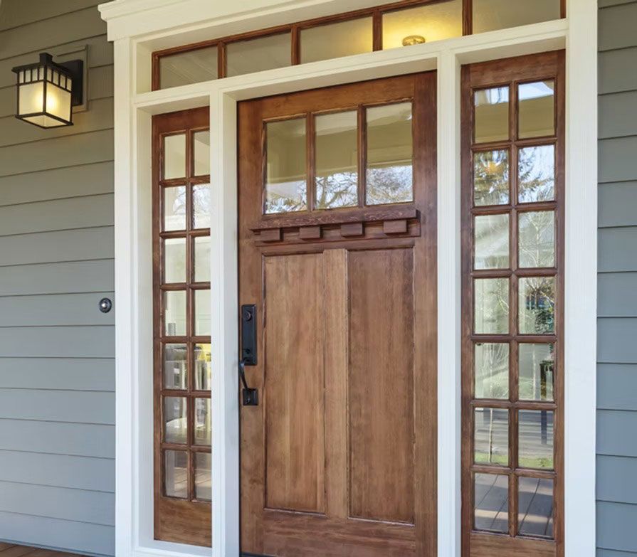 A wood front door with a transom and side windows, framed in white, set against light gray siding with an outdoor lantern.