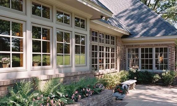 A brick house exterior with large, grid-paned windows, a stone patio, and lush green landscaping.