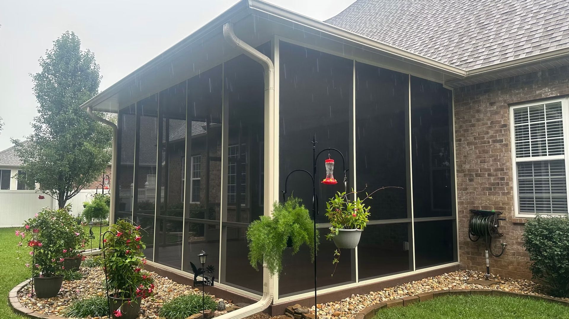 A screened-in patio attached to a brick house, featuring a white frame, a downspout, and hanging plants in a yard.