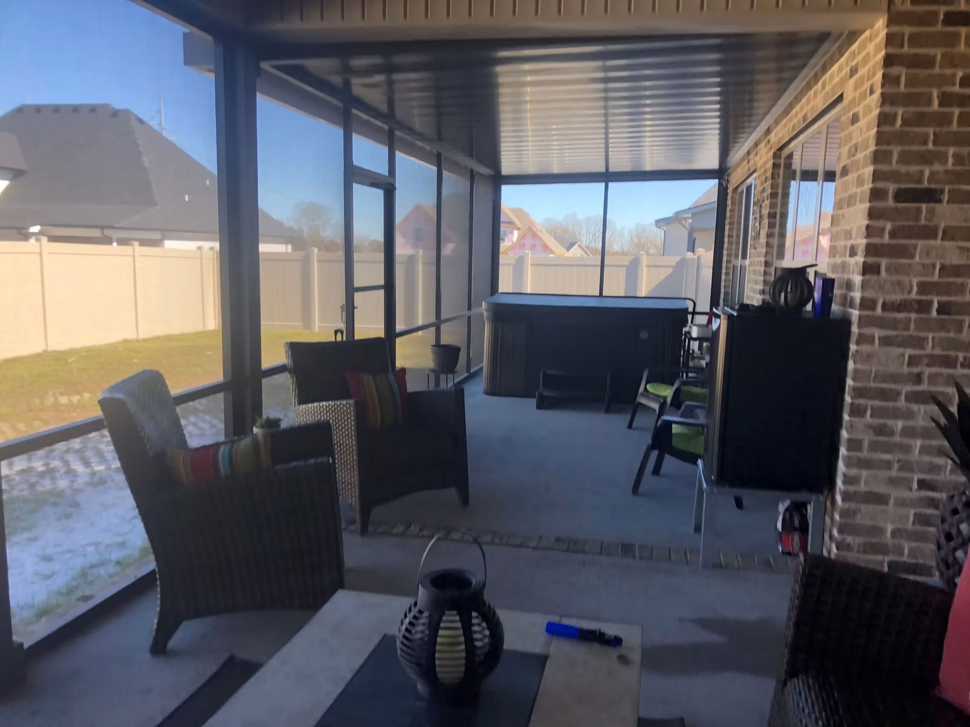 A screened-in patio featuring wicker chairs, a table, and a hot tub under a covered roof in a residential backyard.