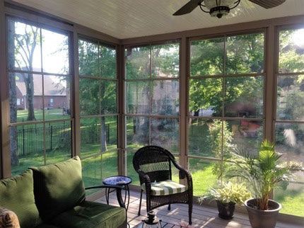A sunroom with green cushions, a wicker chair, a small table, and a potted plant, looking out onto a grassy yard.