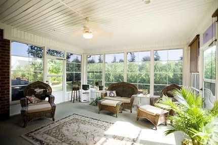 A sunroom with a ceiling fan, wicker furniture, and large windows overlooking lush green trees.